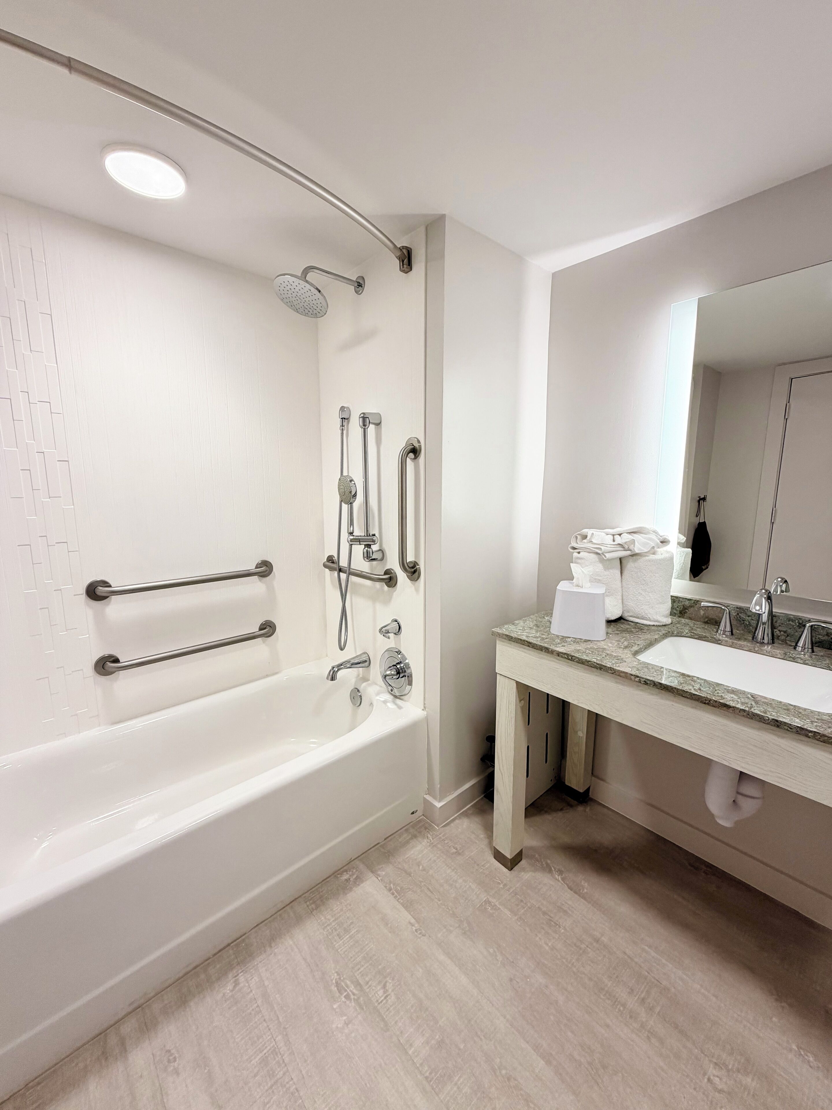 Un baño moderno con una bañera blanca, barras de apoyo, ducha, lavabo con espejo y dispensador de toallas.