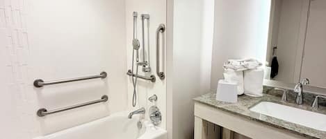 Un baño moderno con una bañera blanca, barras de apoyo, ducha, lavabo con espejo y dispensador de toallas.