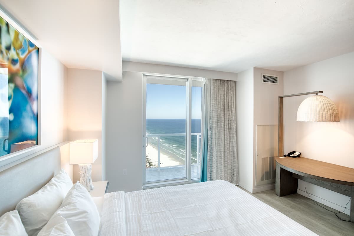 Ein Hotelzimmer mit Bett, Schreibtisch, Lampe, einem Gemälde und Blick auf den Strand.