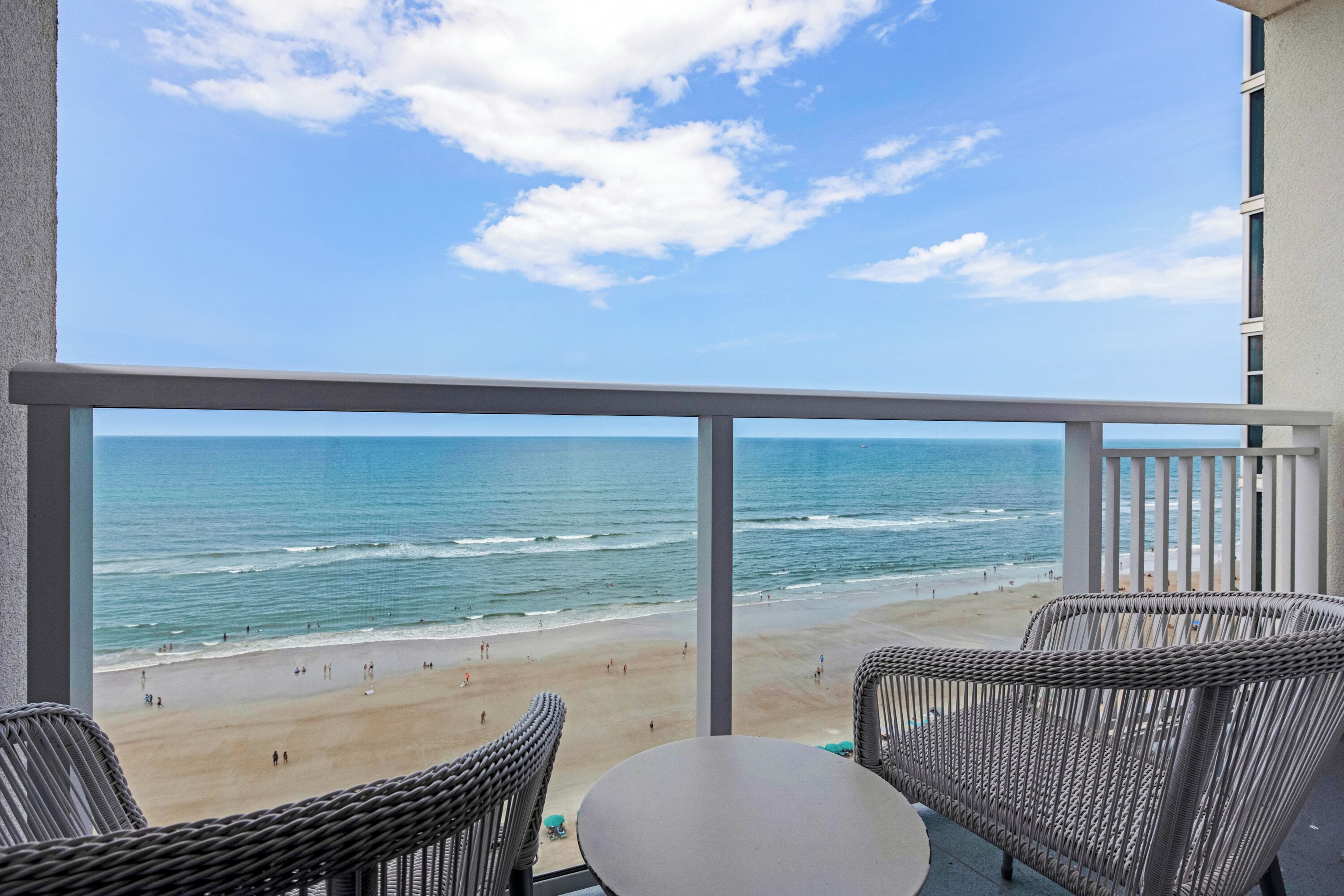 A balcony with wicker chairs and a table overlooking a beach with waves and people.
