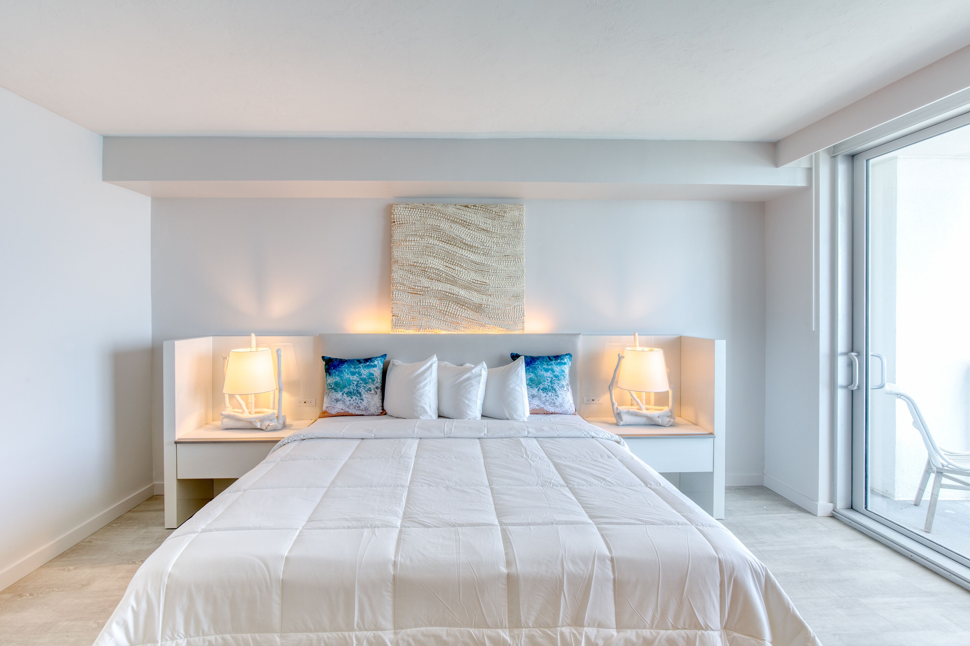 A modern bedroom with a large bed, two bedside tables with lamps, a wall-mounted artwork, and a large window with a view.