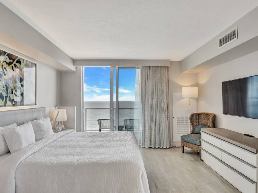 A hotel room with a large bed, a TV, a dresser, a chair, and a balcony with a view.