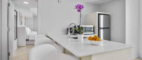 Cocina moderna con encimeras blancas, refrigerador de acero inoxidable, un lavamanos y una orquídea en maceta. Hay un cuenco con fruta sobre la encimera y una silla en primer plano.