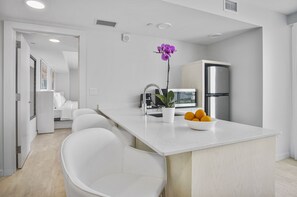 A modern kitchen with white countertops, a stainless steel refrigerator, a sink, and a potted orchid. There is a bowl of fruit on the counter and a chair in the foreground.