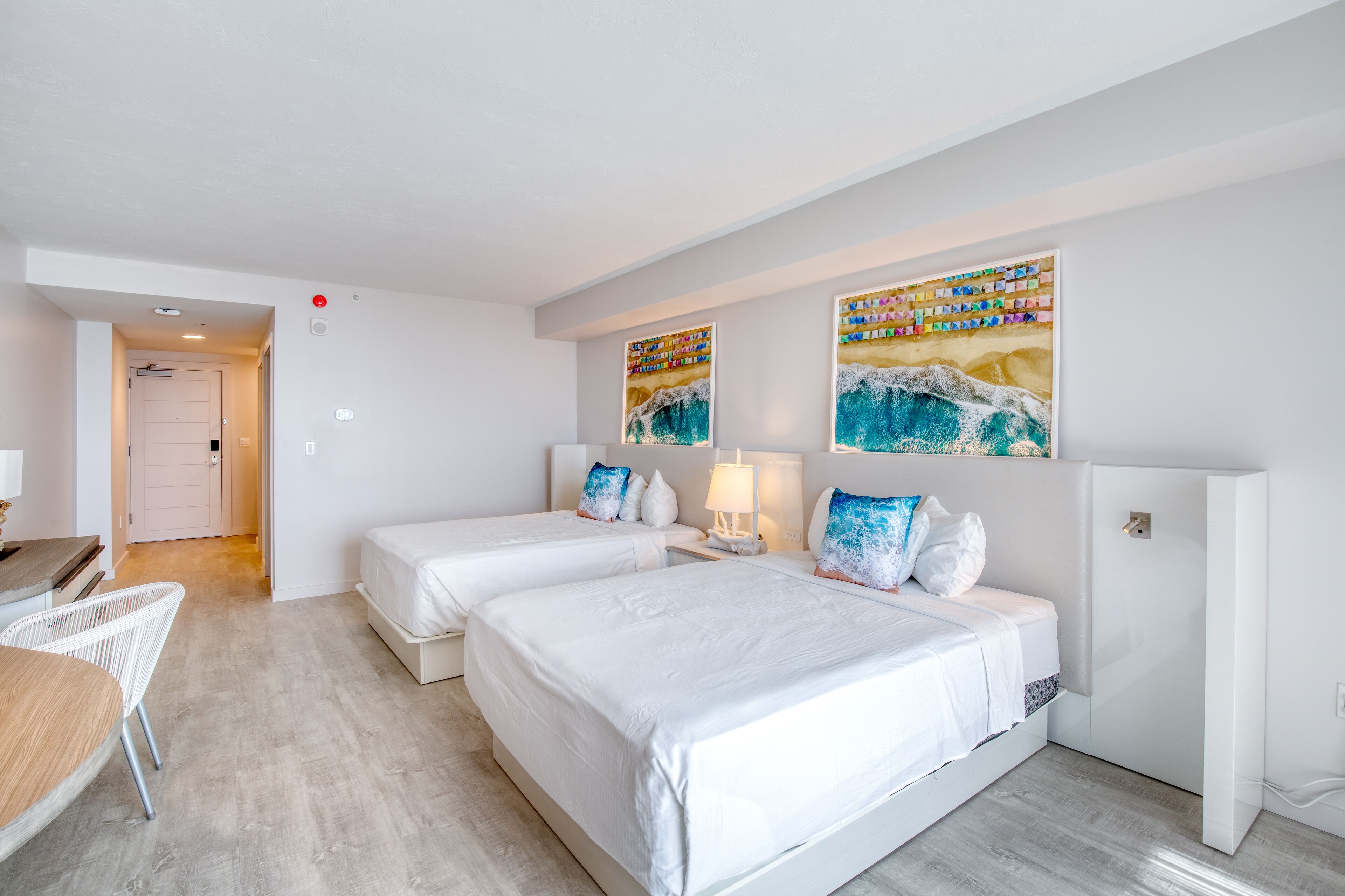 Habitación de hotel con dos camas, un escritorio y una silla. Hay cuadros en la pared y una puerta al fondo.