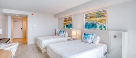 Habitación de hotel con dos camas, un escritorio y una silla. Hay cuadros en la pared y una puerta al fondo.