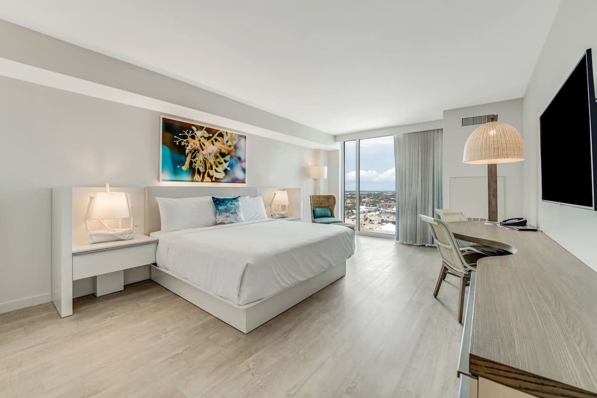 Ein modernes Hotelzimmer mit einem großen Bett, Nachttischlampen, einem Schreibtisch und einem großen Fenster mit Blick auf die Stadt.