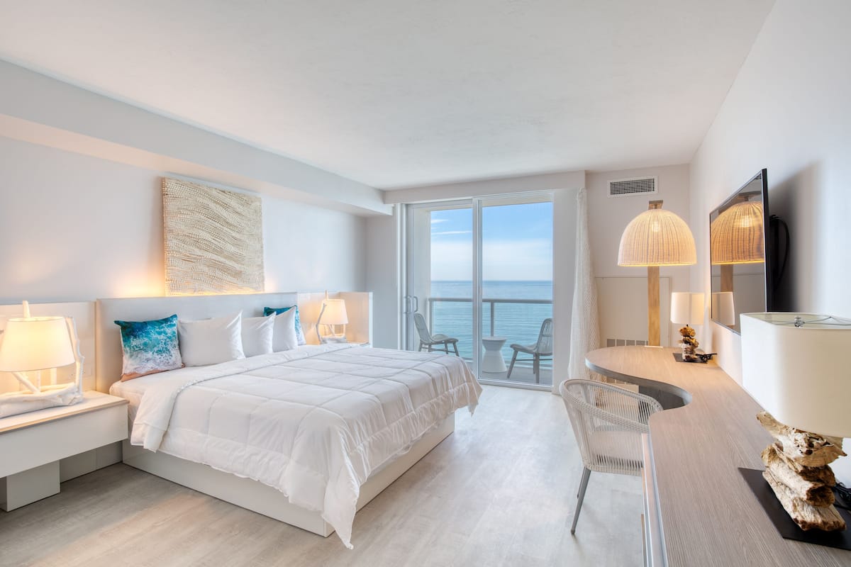 Ein modernes Hotelzimmer mit einem großen Bett, zwei Nachttischen mit Lampen, einem Schreibtisch mit Stuhl und einem großen Fenster mit Meerblick.