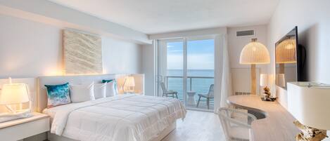 A modern hotel room with a large bed, two bedside tables with lamps, a desk with a chair, and a large window offering a view of the sea.