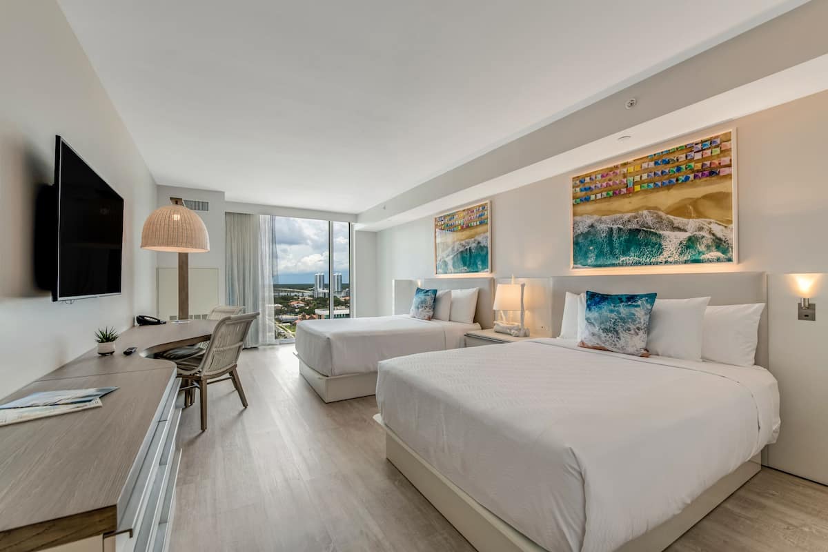 Ein Hotelzimmer mit zwei Betten, einem Schreibtisch und einem großen Fenster mit Blick auf die Stadt.