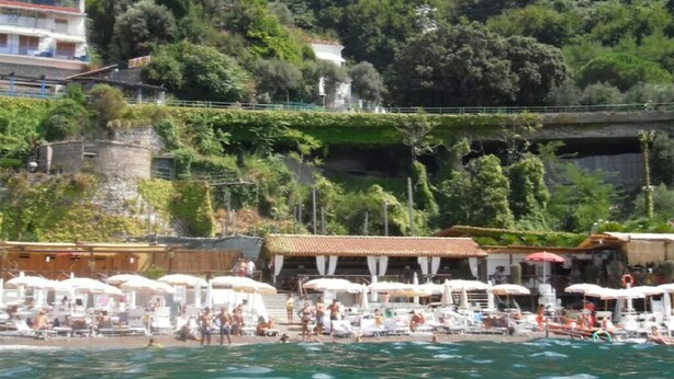 Ein Strand mit Liegestühlen und Sonnenschirmen, ein Gebäude mit rotem Dach und üppiges Grün im Hintergrund.