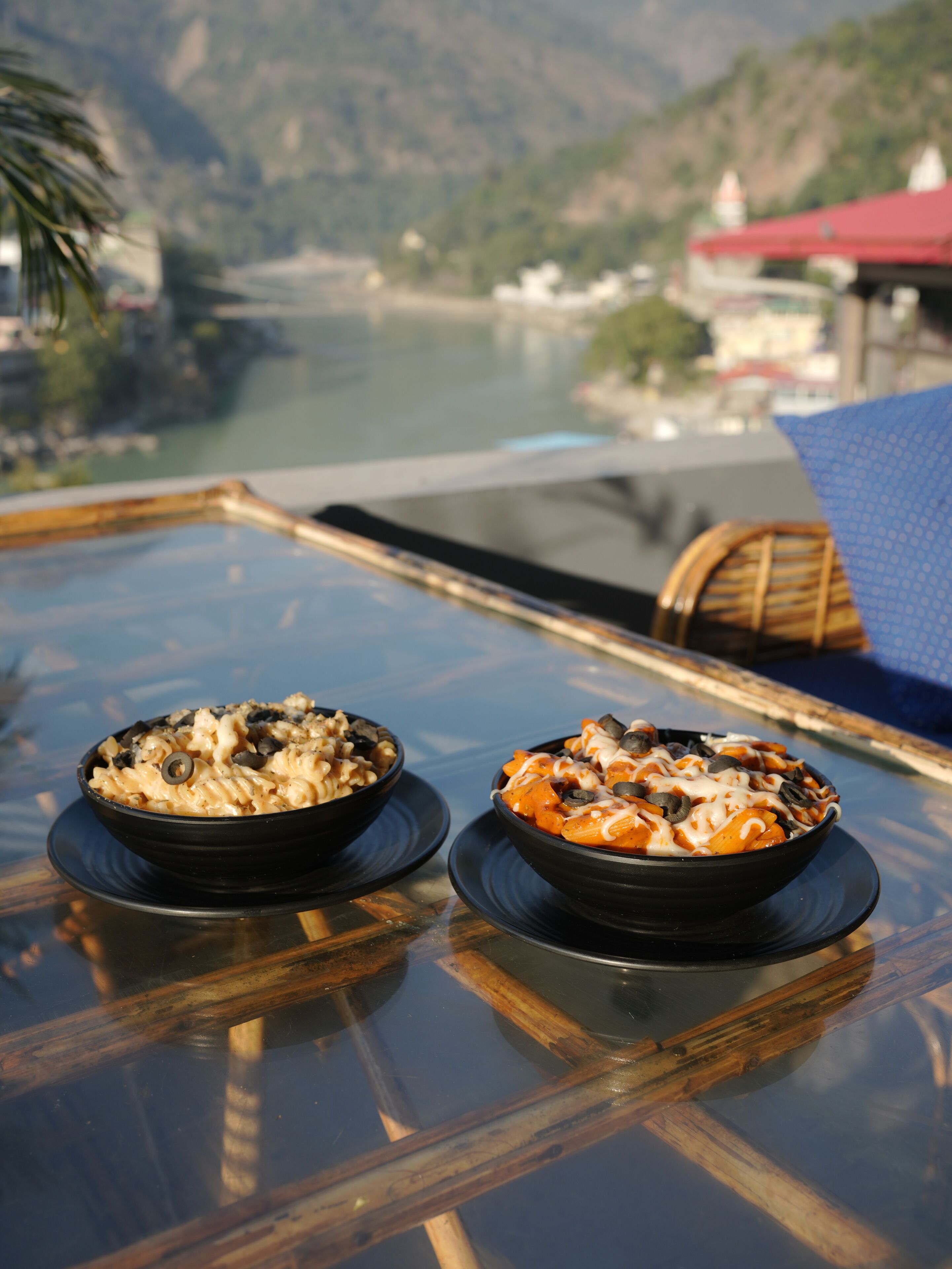 Two bowls of pasta on a glass table with a view of a river and mountains in the background.