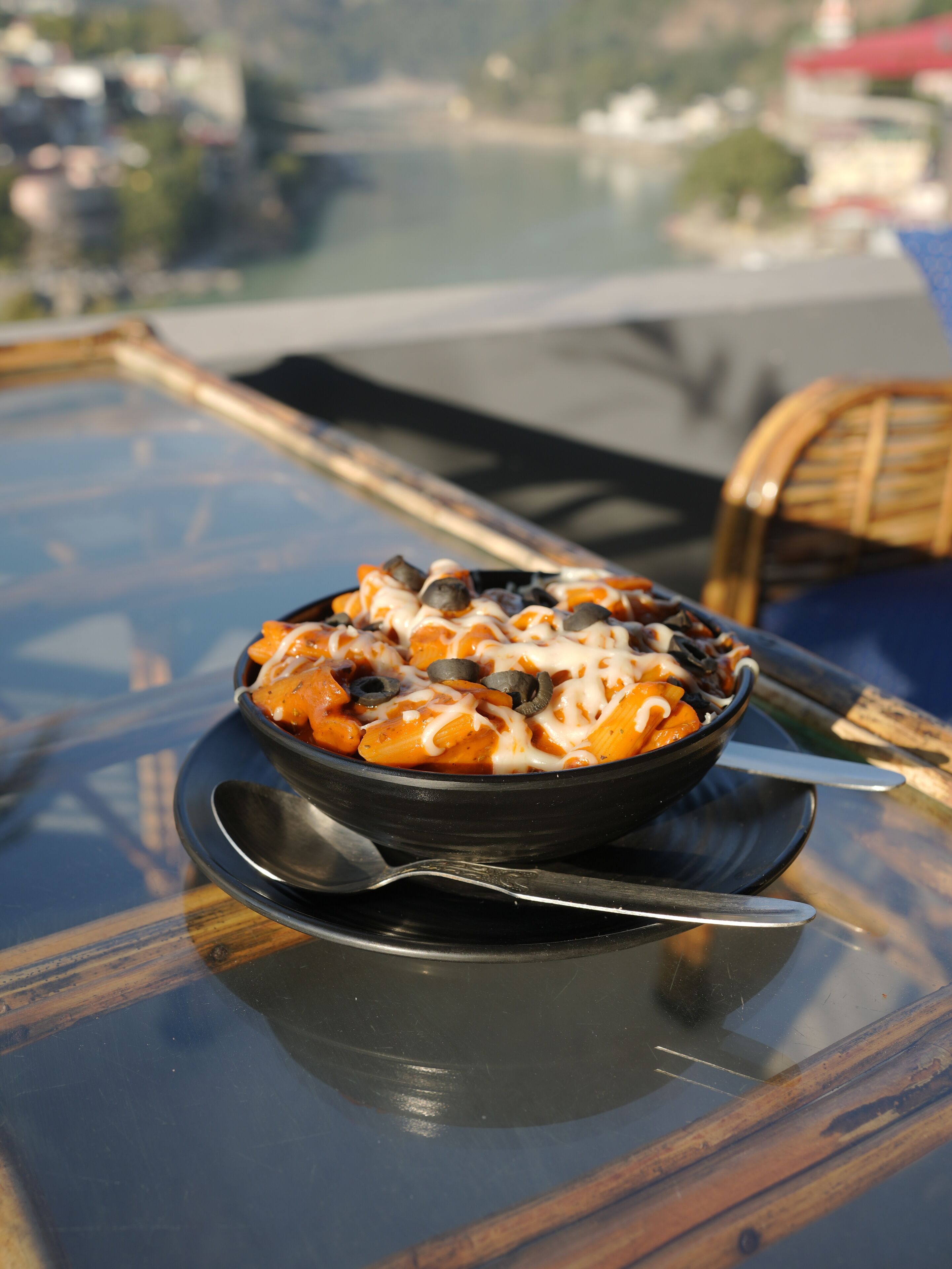A bowl of pasta with a spoon on a glass table with a view of a cityscape in the background.