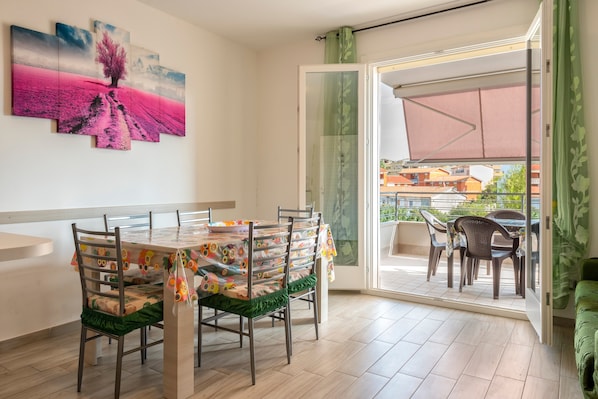 Un coin repas avec une table dressée pour quatre personnes, des chaises avec des coussins verts, un grand tableau accroché au mur et, par une porte-fenêtre coulissante, une vue sur un espace de détente extérieur.