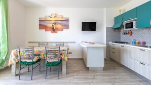 Une cuisine moderne avec des meubles de couleur turquoise, un téléviseur à écran plat et un coin repas agrémenté d’une nappe colorée.
