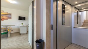 Un couloir intérieur avec un escalier, une poubelle, un téléviseur fixé au mur et un petit bureau agrémenté d’une plante.