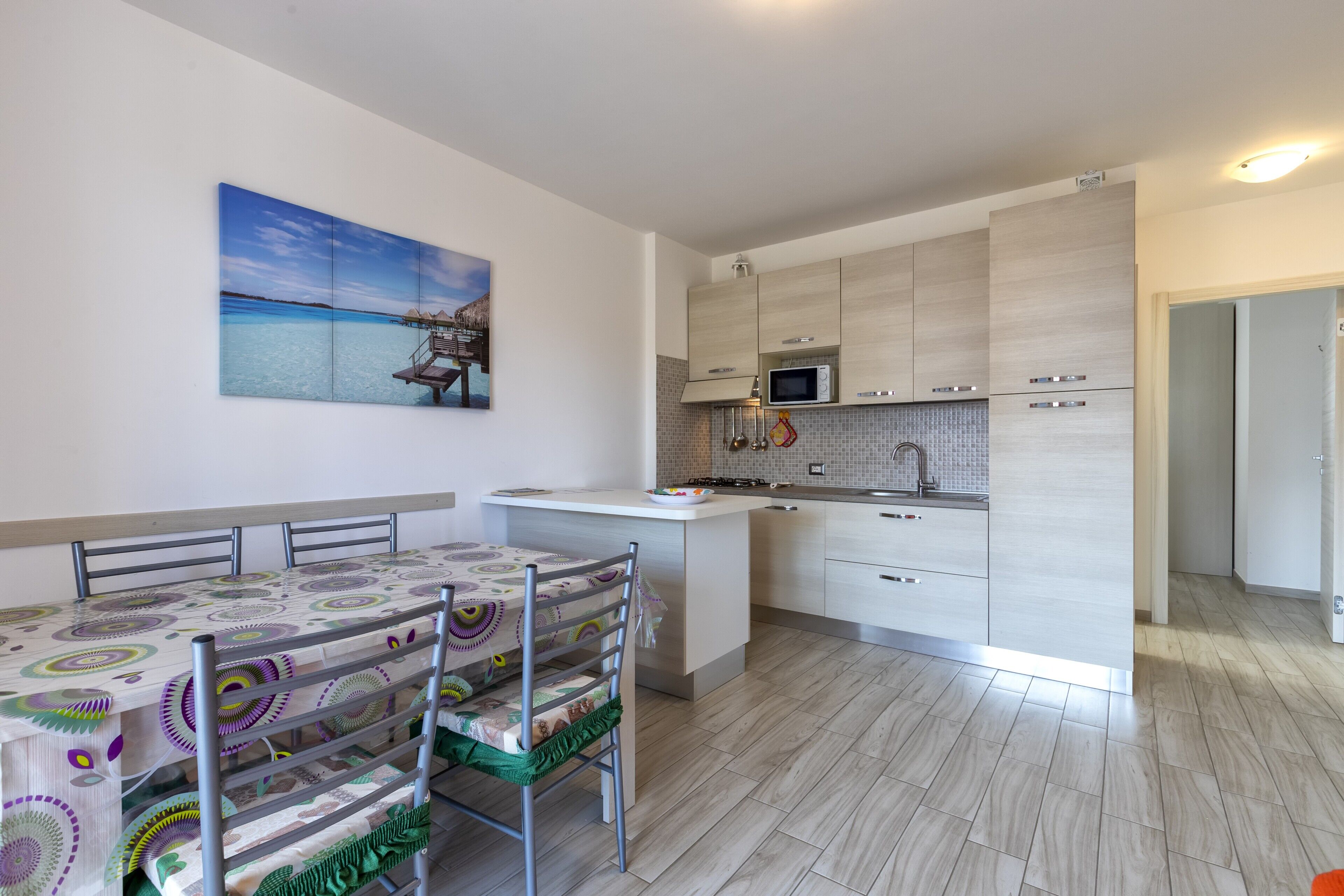 Una cocina moderna con microondas, estufa y armarios, una mesa de comedor con sillas, y un cuadro con una escena de playa.