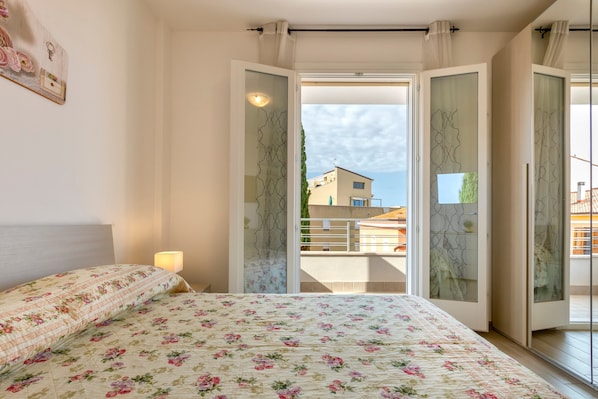 A bedroom with a floral bedspread, a bedside lamp, and a view of a balcony with a building in the background.