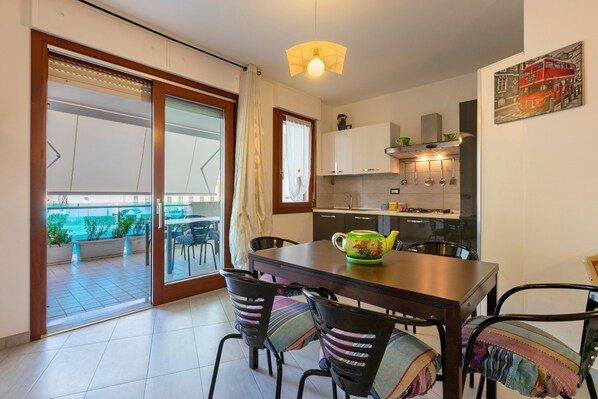Un espace repas comprenant une table, des chaises, une cuisine en arrière-plan et, par une porte coulissante, une vue sur un balcon extérieur.