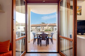 An open sliding door leading to a balcony with a table and chairs, a sofa, and a TV on a stand. - Comfortable Apartment “Egeo” close to the Beach with A/C & Terrace; Parking Available (alghero)