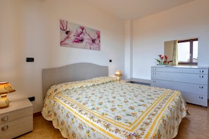 Une chambre à coucher comprenant un lit, une commode, une table de chevet, une lampe, un tableau représentant des fleurs et une fenêtre.
