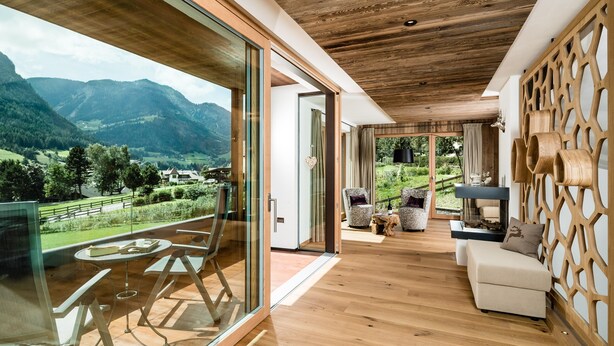 A modern interior with a wooden ceiling, glass sliding doors, a balcony with a table and chairs, and a view of mountains.