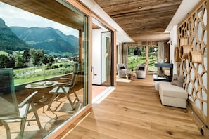 A modern interior with a wooden ceiling, glass sliding doors, a balcony with a table and chairs, and a view of mountains.