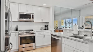 A modern kitchen with white cabinetry, stainless steel appliances, and a large window offering a view of the outdoors. - Gulf Shores Beachfront Condo  Gorgeous views and only steps to the sand! (Holmes Beach)