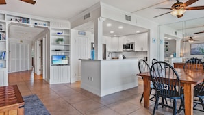 A modern kitchen with white cabinetry, a central island, and a dining area with a wooden table and chairs. - Gulf Shores Beachfront Condo  Gorgeous views and only steps to the sand! (Holmes Beach)