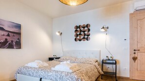 Ein Doppelbett mit gemustertem Überwurf, zwei Nachttische mit Lampen, eine Wandleuchte, eine Holztür und ein gerahmtes Bild an der Wand.