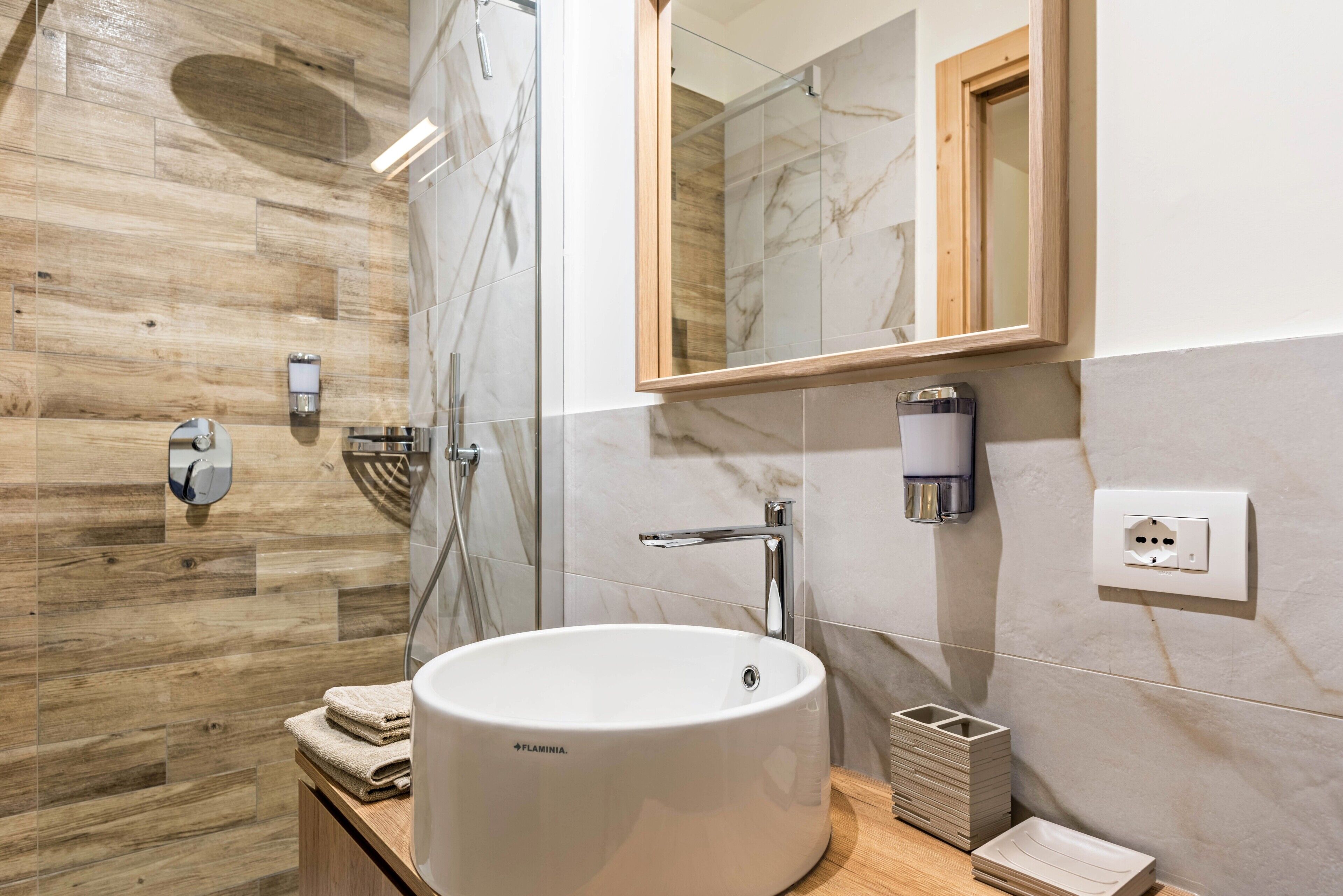 Un baño con piso de madera, un lavamanos blanco, un espejo, un perchero para toallas y una zona de ducha.