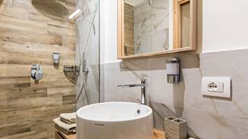 Un baño con piso de madera, un lavamanos blanco, un espejo, un perchero para toallas y una zona de ducha.