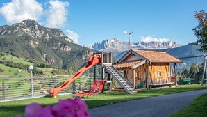 A wooden cabin with a red slide and stairs, surrounded by a metal fence, with a mountain backdrop. - Schlern – Authentic Farm Stay with Animals, Sauna & Mountain Views (Collepietra)
