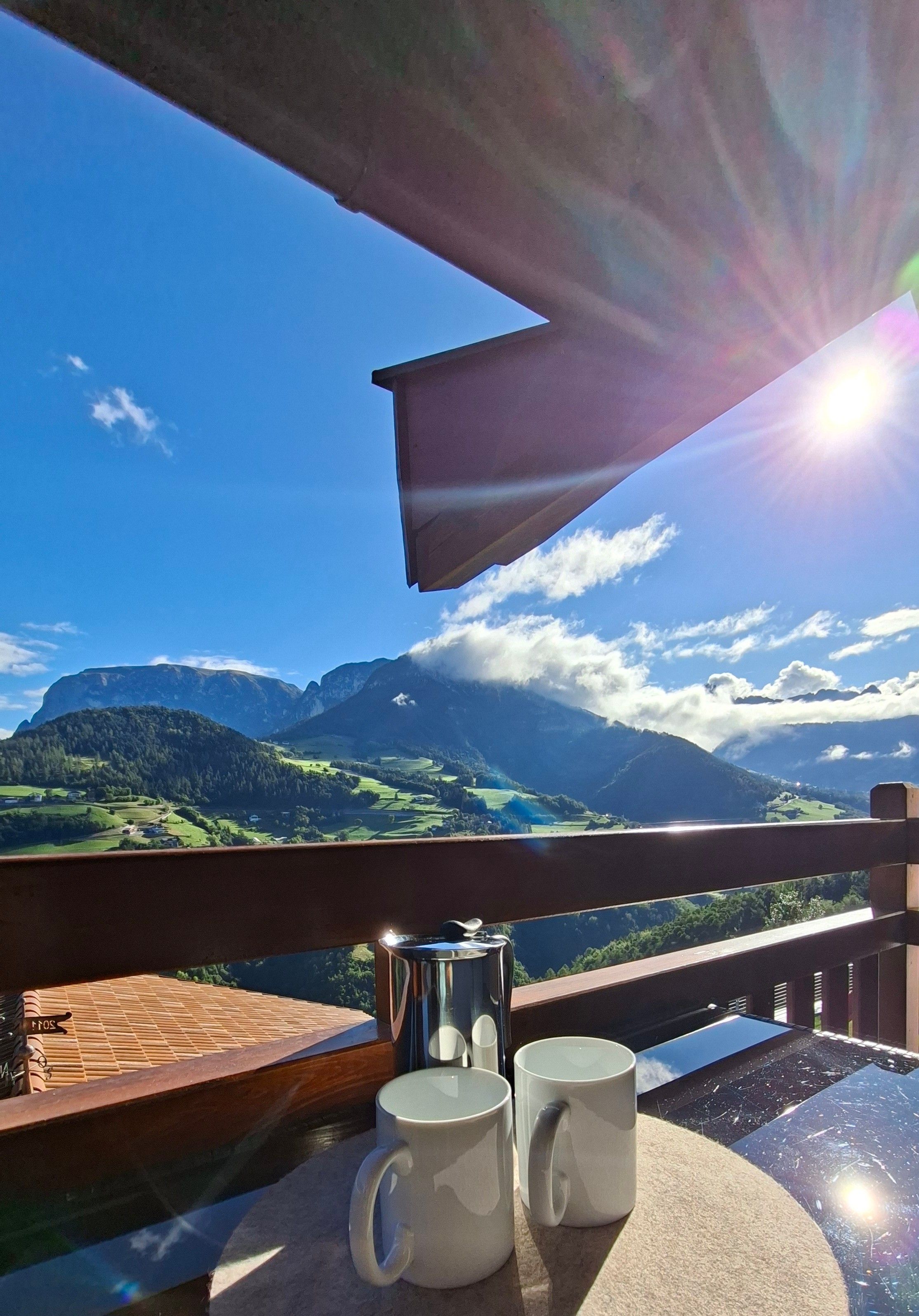 A balcony with a view of mountains, a table with two mugs, and a coffee pot.