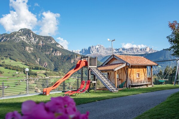 Une cabane en bois avec un toboggan rouge et des escaliers, entourée d’une clôture en métal, avec une montagne en arrière-plan.