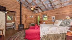 A rustic wooden room with a bed, a red chair, a small table, and a ceiling fan. - Ada's Whole House 3 Bedroom Apts by RedAwning (Fredericksburg)