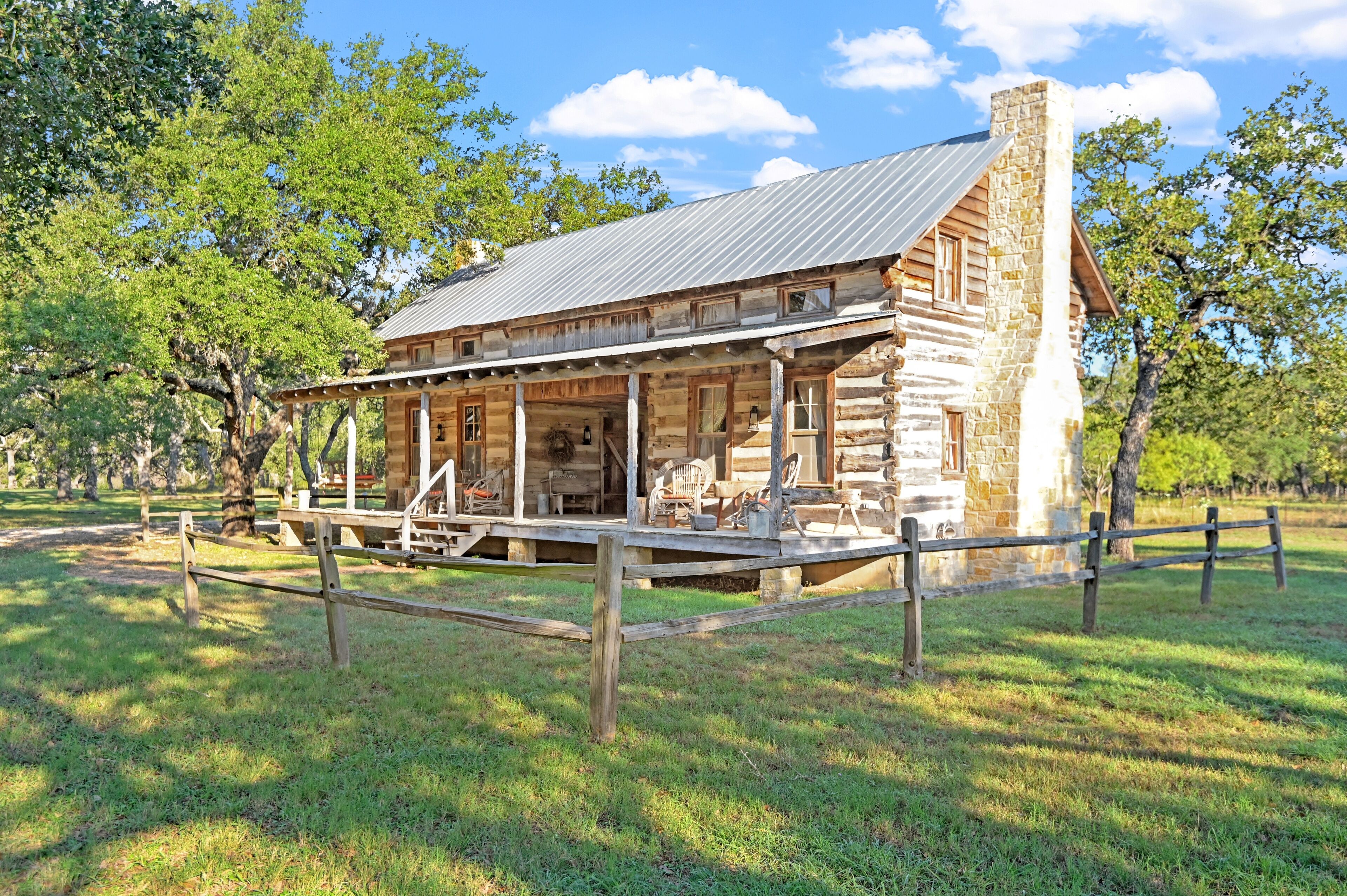 A rustic log cabin with a porch and a chimney, surrounded by trees and a wooden fence.