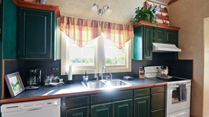 Une cuisine avec des meubles de couleur verte, un lave-vaisselle blanc, une cuisinière et une fenêtre agrémentée d’une lambrequin à rayures.