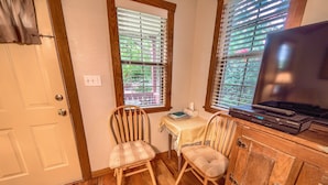 A small room with a wooden floor, a white door, two windows with blinds, a table with a book, a chair, and a TV on a wooden cabinet. - Waldrip Studio Bedroom Cabin by RedAwning (Fredericksburg)