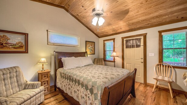 A bedroom with a wooden ceiling, a bed with a floral bedspread, a chair, a nightstand with a lamp, and a window with blinds. - Waldrip Studio Bedroom Cabin by RedAwning (Fredericksburg)
