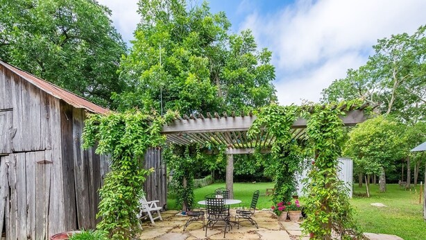 Eine rustikale Terrasse mit Steinbelag, einer mit Grünpflanzen bewachsenen Holzpergola und einem Holzstall im Hintergrund.
