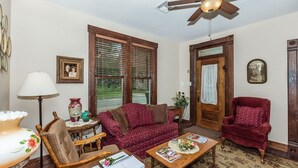 Ein gemütliches Wohnzimmer mit Deckenventilator aus Holz, einem roten Samt-Sessel, einem gemusterten Sofa und einem Couchtisch mit Zeitschriften und einer Vase.