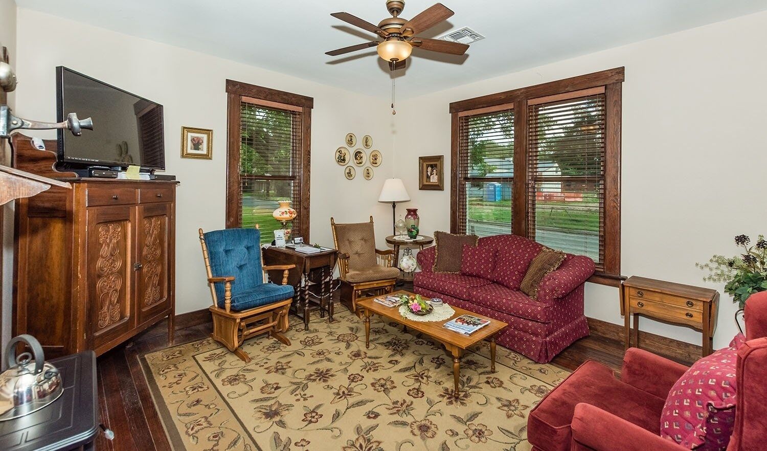 A living room with a wooden cabinet, a television, a ceiling fan, a red sofa, a blue chair, a wooden coffee table, and a patterned rug.