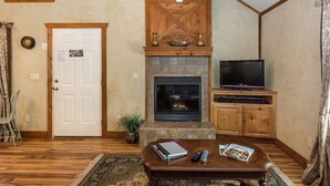 A cozy living room with a fireplace, wooden furniture, a television, and a door with a 'Please Wait' sign. - Appian Way - Oliva Studio Bedroom Cottage by RedAwning (Fredericksburg)
