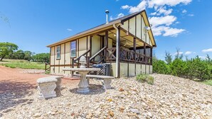 A single-story building with a gabled roof, a chimney, and a covered porch with a table and chairs. - Appian Way - Oliva Studio Bedroom Cottage by RedAwning (Fredericksburg)