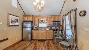 A compact kitchen with wooden cabinets, stainless steel appliances, a chandelier, and a dining area with a table and chairs. - Appian Way - Oliva Studio Bedroom Cottage by RedAwning (Fredericksburg)