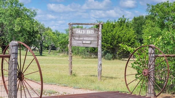 A rustic wooden sign for OMA'S HAUS with a bed and breakfast service, featuring two large, rusted wheels in the foreground. - Oma's Haus 2 Bedroom Home by RedAwning (Fredericksburg)