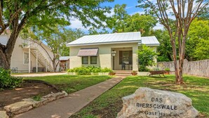 A single-story house with a front porch, a small garden, and a stone marker labeled "DAS BIERSCHWALE Gast Haus 512 WA San Antonio". - Das Bierschwale Gastehaus 2 Bedroom Home by RedAwning (Fredericksburg)