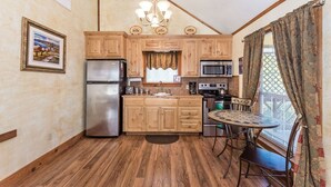 A compact kitchen with wooden cabinets, a refrigerator, a microwave, and a stove. There is a dining table with chairs and a window with curtains.