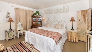 A bedroom with a large bed, a wooden cabinet, a white chair, a small table, and a decorative wall piece. - Binky's Summer Afternoon Studio Bedroom Cottage by RedAwning (Fredericksburg)